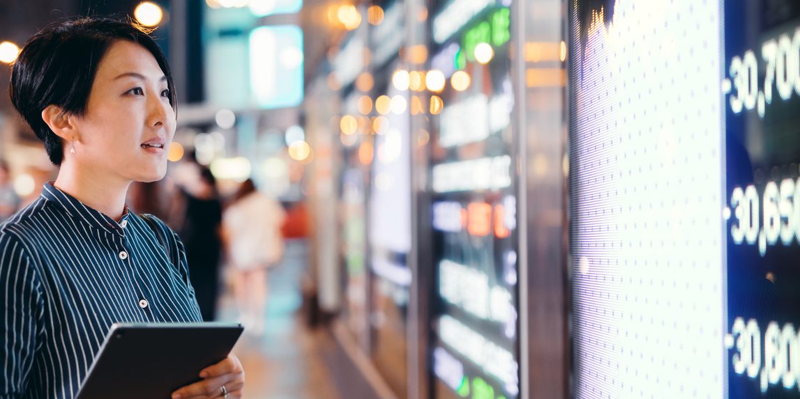 Asian businesswomen checking stock market data on tablet before Hong Kong financial display board