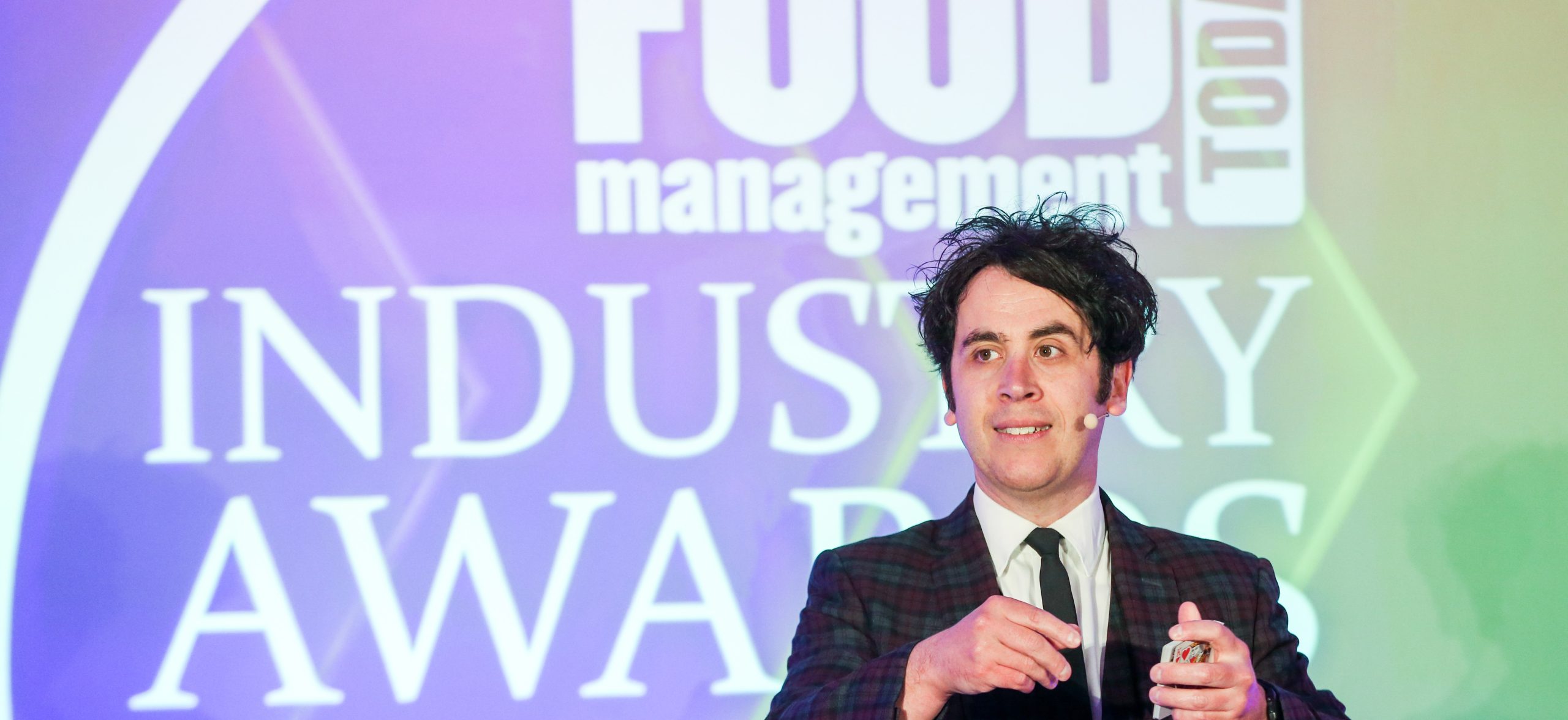 Pete Firman stands offset to the right on a stage in front of a screen that reads 'Food Management Today Industry Awards'. He holds a deck of cards in his hands.